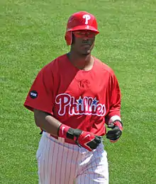 A dark-skinned young man wearing a red baseball jersey and batting helmet and white pinstriped baseball pants