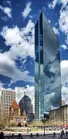 Ground-level view of a 60-story skyscraper; the clouds in the sky are reflected off the building's tinted, all-glass facade. On the ground to the left of the building is a Romanesque church, surrounded by a grassy public square. Two shorter skyscrapers are visible in the distance.