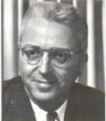 A smiling white man with white hair, wearing glasses and a suit