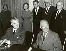 Diefenbaker and a smiling bald man in a suit sit at a table.  Two women and two men stand behind them.