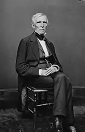 A man with gray hair wearing a black suit and white shirt, sitting with his hands folded