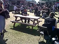 TT rider John McGuinness awaiting a press interview at a picnic bench in the public catering area at the rear of the TT Grandstand during 2013 TT races, with Mrs McGuinness standing to left