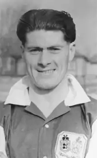 Monochrome head-and-shoulders photo of young white man with dark hair brushed straight back. He is wearing an open-necked shirt with white collar and sleeves. The shirt has a large badge on the left breast.