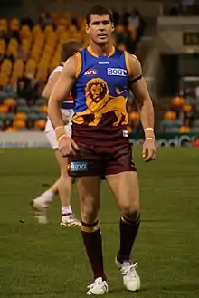 A dark-brown-haired footballer in a sleeveless blue-and-maroon guernsey emblazoned with a golden lion standing on a grassed oval