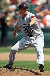 A man prepares to deliver a baseball pitch, holding the ball behind his body with his right hand. He is wearing a gray baseball uniform with "Boston" across the chest in navy letters and a navy blue baseball cap with a red "B". A black baseball glove is on his left hand, held in front of his body as he strides forward.