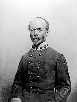 Black and white photograph shows a balding man with a salt-and-pepper moustache and beard. He wears a double-breasted gray military uniform with three stars on the collar.