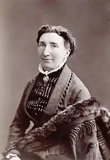 A white woman, wearing a high-collared bodice, a cameo broach at the throat, and a fur-trimmed cloak