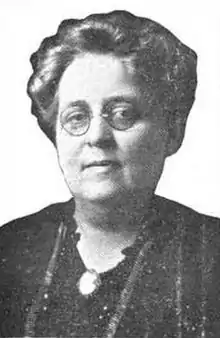 A middle-aged white woman with short grey hair, wearing round glasses and a dark blouse or dress