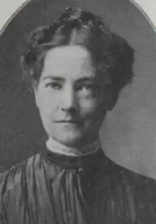 A white woman with dark hair in an updo, wearing a high-collared dark dress