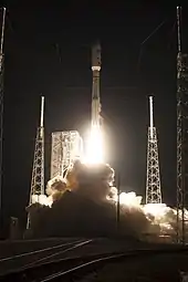 A view close to the launch pad of a rocket shortly after lift-off, along with four structures associated with the lightning protection system and the service structure partially obscured by the rocket's exhaust.