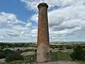 Large cylindrical red brick chimney, cut earth, trees and countryside in background