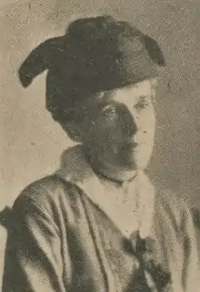 An older woman wearing a dark hat, and a dress with a white collar. she has a pendant on a choker at her neck.