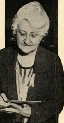 A white woman with white hair, gaze cast on a book in her hands; she is wearing a jacket and a soft blouse with a scooped neckline