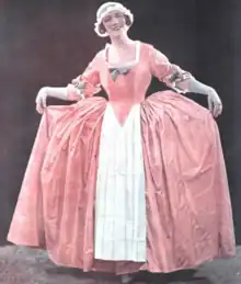 A smiling white woman, standing on stage, wearing an 18th century-style costume, a wide pink dress with a white apron and cap