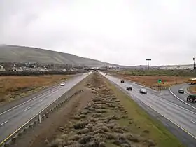 A four-lane freeway with a wide median approaching a residential neighborhood with an overpass in the distance.