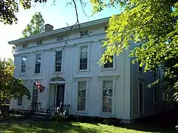 A white house seen from slightly to its right, with a tree branch partially obscuring it. Its front windows and door have some decorative features like columns. An American flag flies from a pole above the main entrance.