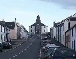 A wide street leads up a slope with parked cars and stone houses painted in whites and yellows on either side. At the end of the street there is a grey and white building with a short spire.