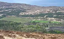 A view from an elevation above cultivated fields and a mixed wood of conifers and deciduous trees. Various indistinct buildings are visible among these trees. The brown ridge of a hill in the background has conifers growing on its lower slopes.