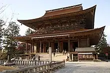 Large wooden building with a hip-and-gable roof and an enclosing pent roof. There is a railed open veranda above the pent roof.