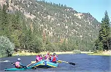 A total of five people, four in an inflatable raft and one in a kayak, floating down a calm section of blue water