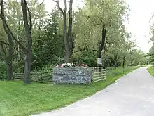 A narrow paved road on the right disappears into the distance. It is lined with trees, and the foreground shows a mown lawn fronting an open gate, with a wooden fence leading away to the right enclosing the forest behind it.