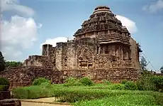 Picture of a square-shaped stone temple, with straight walls and a conical top. In front of the temple, stone structures are seen, as well as a green garden.