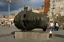 In an open plaza, a large, hollow head made of dark metal with empty eye sockets lies on its side on a concrete slab.