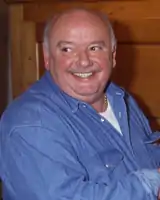 A man in his fifties, balding and portly, is shown smiling to the left of the camera.