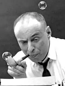 A black-and-white photo of a bald middle-aged man, blowing bubbles from a bubble pipe