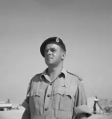 A black and white photograph of the head and shoulders of a man in a military uniform and beret. In the background is a tent and military vehicle