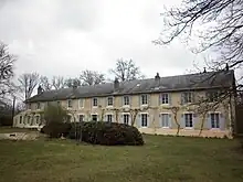 Photograph of the main building of the Colonie, Condé-sur-Vesgre. It is a long symmetrical one-storey building with large regular windows, six on each side of the door. In front of it is a small grassy area with bushes and a few trees.