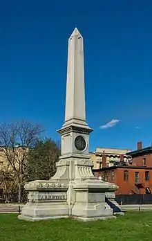 Picture of a white stone monument standing on green grass with a blue sky in the background.