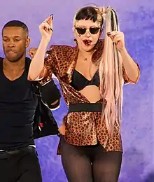 A blond woman wearing black sunglass and her hair in a side ponytail. She wears a black and gold printed shirt through which her bustier is seen. The woman wears black stockings and has both of her hands raised a little upwards. She is flanked on her right by man, who strikes a similar pose, wearing black vest and pants. They are standing in front of a light blue backdrop.