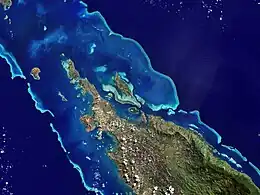 Satellite image of the tip of a large island fringed by barrier reefs, with different hues of blue showing the immediate difference in water depth created by the reefs