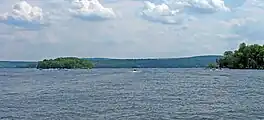 Image of a boat moving away from the photographer on a wide lake with two forested islands visible in the foreground. Mountains rise in the background.