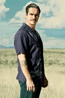 A man stands in the desert in broad daylight, looking at the camera in a slightly intimidating manner