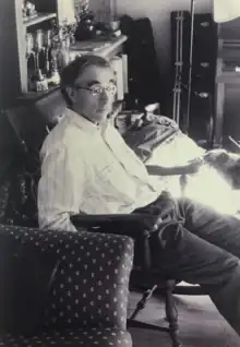 A black and white photo depicting a middle-aged man, Larry Gordon, sitting in a carved wooden chair in a relaxed posture, wearing wire-frame glasses, a light-coloured Oxford shirt, and dark-coloured trousers. Behind him are a series of musical instruments in cases. The room appears warm and sunny.