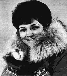 Portrait of a Native American woman with short hair wearing a parka with fur around the neck and decorative cuffs which are visible because her hands are raised to her chin.