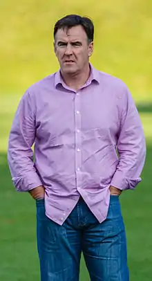 A brown-haired man with blue jeans and a purple shirt stands on the grass of a football field.