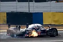 A photograph of a sports prototype racing car with flames coming out of its right-rear corner