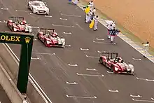 Four racing cars being driven on a motor racing circuit with people to their left waving flags and applauding them in congratulations.