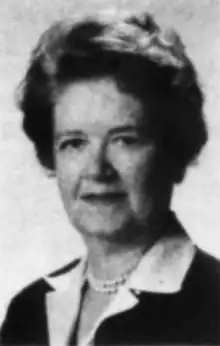 A middle-aged white woman with short dark coiffed hair, wearing a dark suit, white blouse, and pearls