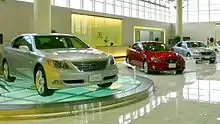 Car showroom displaying three sedans, the nearest on a glass turntable, in front of a reception counter and windows.