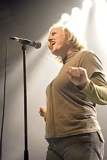 A Caucasian female with shoulder-length blond hair singing into a microphone