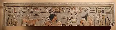 Light gray stone surface with carved and painted images of two woman, a falcon-headed god, a black-haired man with a long goatee, a jackal-headed god, and Egyptian hieroglyphs inscribed along the top