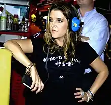 A woman in a black T-shirt holds her left hand on her hip, while leaning against a chest-height surface with her right elbow. She is wearing blue headphones and looks towards her right.