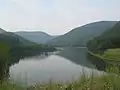 View from atop a grassy dam of a lake between and reflecting green mountains