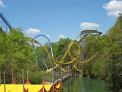 The Loch Ness Monster is viewed from a vantage point on an elevated surface overlooking the park's lake. On the bottom left, a tent structure is viewed with a footbridge across the lake centered. The Loch Ness Monster takes up the center image with a train about to descend the first drop. The Griffon Dive Coaster is also seen in the background.