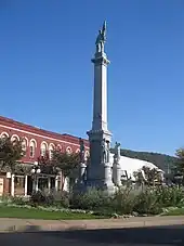 A stone monument about three stories tall rises from a park-like area along a city street. The monument features stone replicas of uniformed soldiers.