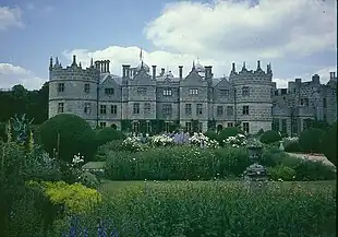 Castle of three stories, in stone, with two towers with five bays between them, the second and fourth bay protruding slightly. It has ornate gables and a flower garden is in the foreground.